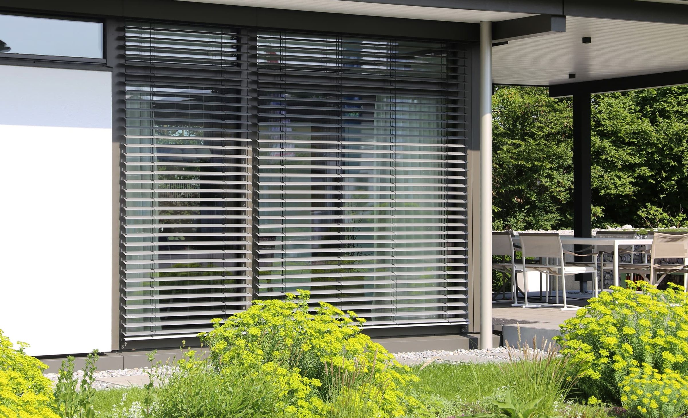 Moderne Hausfassade mit anthrazitfarbenen Außen-Raffstores vor einer Terrasse mit Gartenbepflanzung.